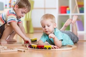 zwei als männlich zu lesende Kleinkinder spielen in einem Zimmer mit einer Holzeisenbahn.