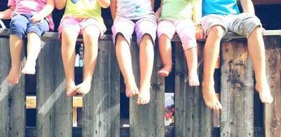 Das Bild zeigt fünf Kinder, die nebeneinander auf einem Holzzaun sitzen und die Füße herabbaumeln lassen. Es ist jedoch nur die untere Hälfte des Körpers der Kinder zu sehen. Die obere Hälfte ist durch den begrenzten Bildausschnitt abgeschnitten.