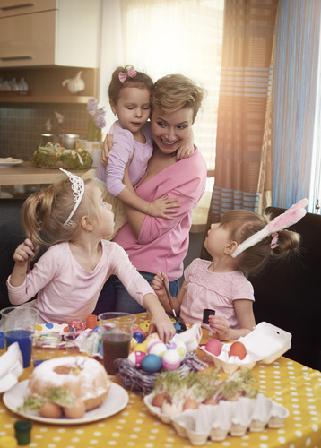 Das Bild zeigt eine Küchen-Esszimmer-Szene an Ostern. Eine blonde kurzhaarige Frau mit rosa Oberteil hält ein weibliches Kleinkind auf dem Arm. Am Esstisch sitzen zwei etwas ältere blondhaarige weibliche rosa gekleidete Kleinkinder, die zu der Frau aufschauen. Das linke Kind trägt ein Prinzessinnendiadem, das rechte Kind Hasenohren als Haarreif auf dem Kopf. Auf dem Tisch mit gelber Tischdecke mit weißen Punkten befinden sich ein Gugelhupfkuchen und bemalte Eier in einem Osternest.